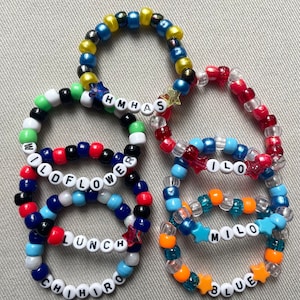 May include: A collection of beaded bracelets in various colors, including blue, red, and orange. Some bracelets spell out words like "WILDFLOWER," "LUNCH," and "MILO" using white letter beads. Star-shaped beads add decorative accents.