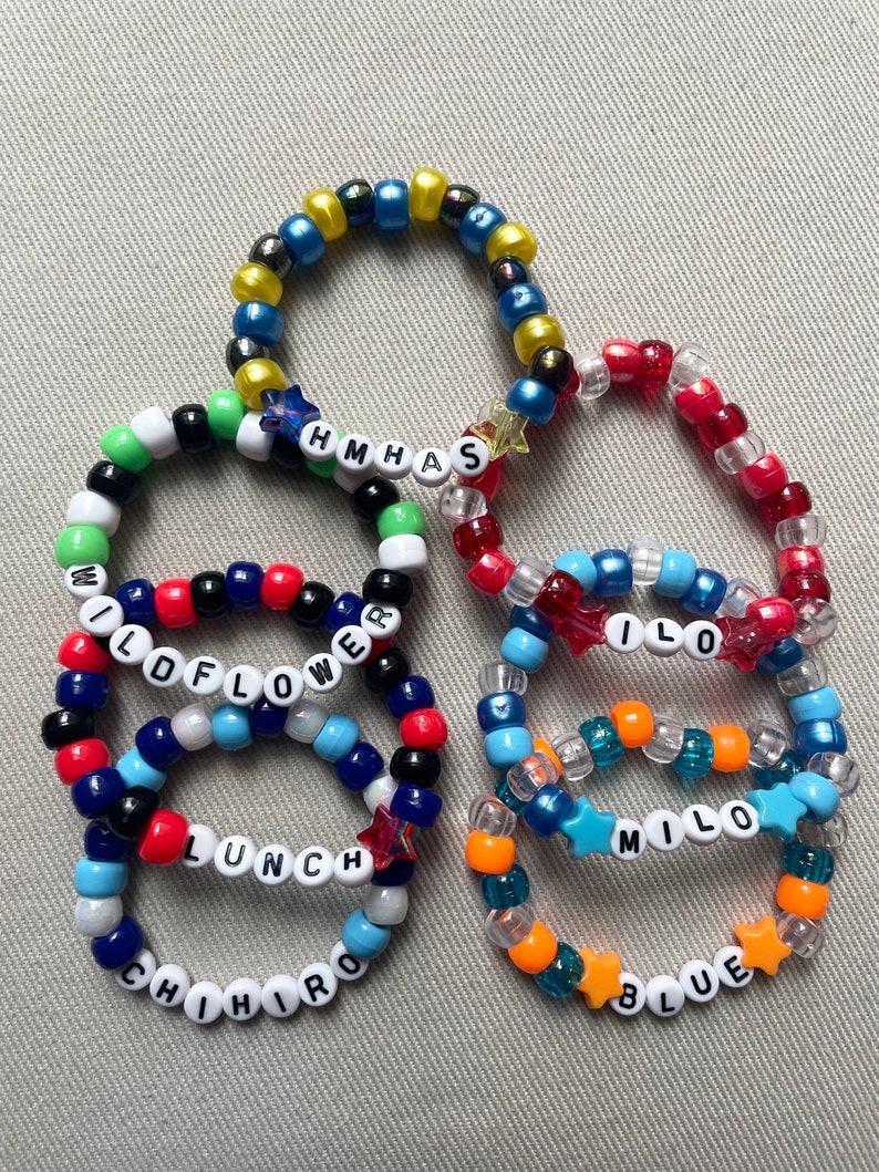 May include: A collection of beaded bracelets in various colors, including blue, red, and orange. Some bracelets spell out words like "WILDFLOWER," "LUNCH," and "MILO" using white letter beads. Star-shaped beads add decorative accents.