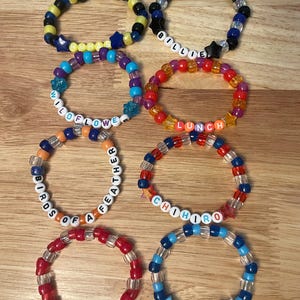 May include: A collection of colorful beaded bracelets with various designs and text. The bracelets feature a mix of colors, including blue, yellow, red, and clear beads. Some bracelets spell out words like "WILDFLOWER" and "LUNCH".