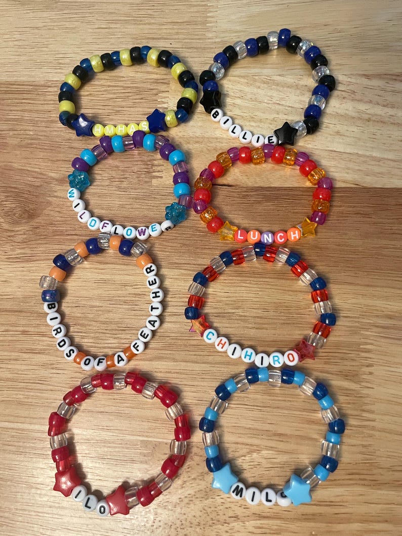 May include: A collection of colorful beaded bracelets with various designs and text. The bracelets feature a mix of colors, including blue, yellow, red, and clear beads. Some bracelets spell out words like "WILDFLOWER" and "LUNCH".