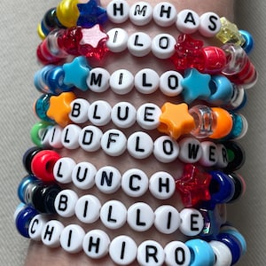 May include: A collection of colorful beaded bracelets. Each bracelet features white letter beads spelling out names and words like "HMHAS", "MILO", "BLUE", "WILDFLOWER", "LUNCH", "BILLIE", and "CHIHIRO". The bracelets include star-shaped and other decorative beads.