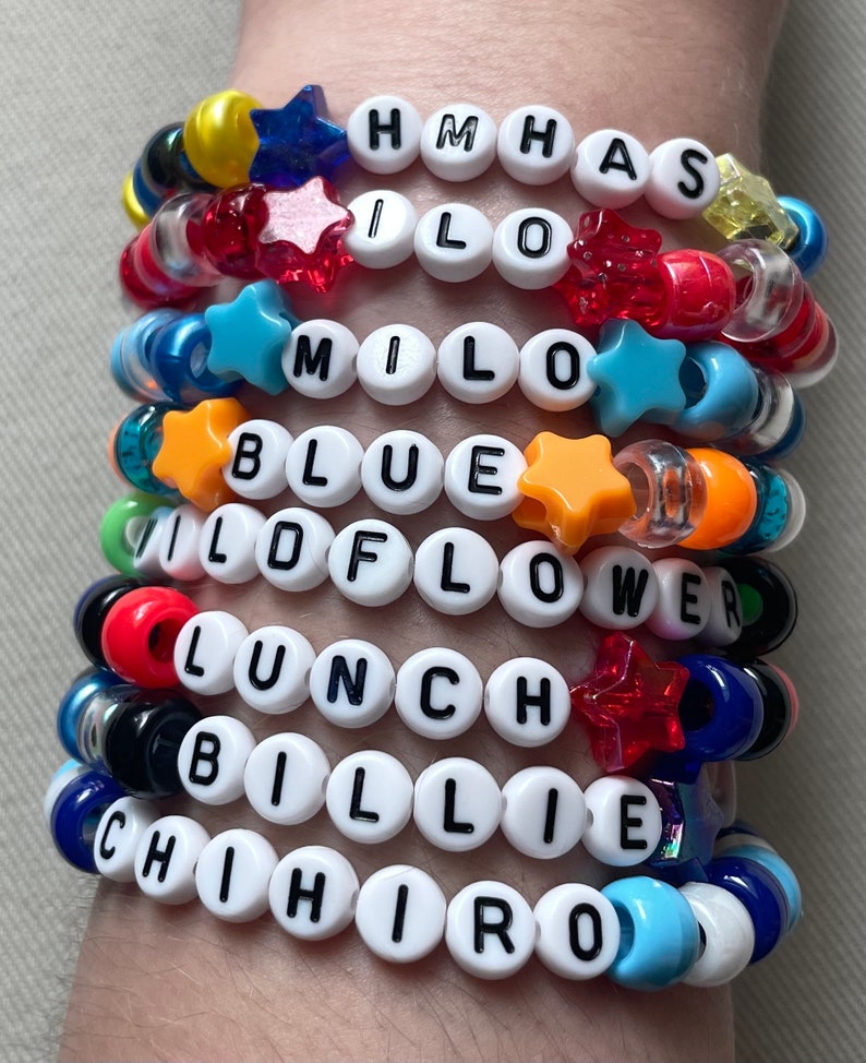 May include: A collection of colorful beaded bracelets. Each bracelet features white letter beads spelling out names and words like "HMHAS", "MILO", "BLUE", "WILDFLOWER", "LUNCH", "BILLIE", and "CHIHIRO". The bracelets include star-shaped and other decorative beads.