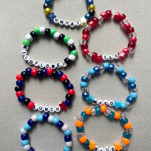 May include: A collection of beaded bracelets in various colors, including blue, red, green, and orange. Some bracelets feature white letter beads spelling out words like "WILDFLOWER", "LUNCH", and "BLUE". Star-shaped beads are also incorporated.