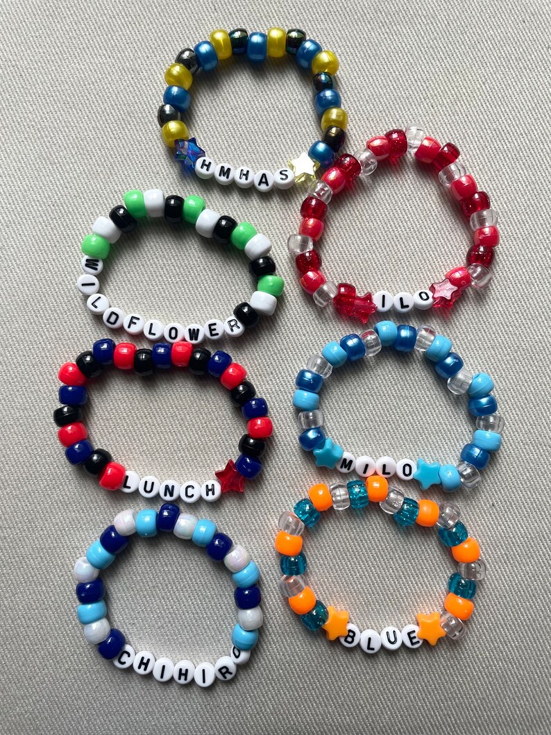 May include: A collection of beaded bracelets in various colors, including blue, red, green, and orange. Some bracelets feature white letter beads spelling out words like "WILDFLOWER", "LUNCH", and "BLUE". Star-shaped beads are also incorporated.