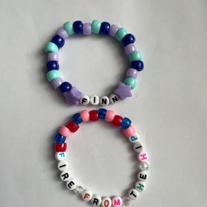 May include: Two beaded bracelets. The top bracelet has blue, purple, and teal beads with the name "FINN" spelled out in white letter beads and purple star-shaped beads. The bottom bracelet has the words "FIRE FROM THE SHIP" spelled out in white letter beads, with red, pink, and blue beads.