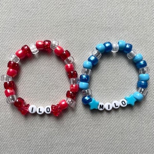May include: Two beaded bracelets. One is red, clear, and white with the name "ILO". The other is blue, clear, and white with the name "MILO". The "MILO" bracelet has star-shaped beads.