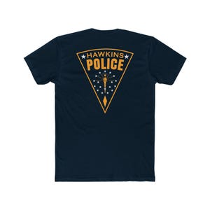 May include: A dark navy blue short-sleeve t-shirt featuring a large triangular graphic on the back. The graphic is outlined in yellow and contains the text HAWKINS POLICE in yellow, along with a central torch icon surrounded by nineteen white stars.