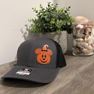 May include: A gray and black trucker hat with a pumpkin-shaped design. The orange pumpkin has mouse ears and a witch hat. The hat has a curved brim and a mesh back. The hat is sitting on a wooden surface.