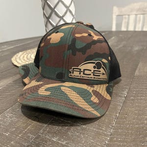May include: A black and green camouflage trucker hat with a black mesh back. The hat has a brown patch with the text "ACE ALPHA CENTAURI EXPEDITIONS" in black.