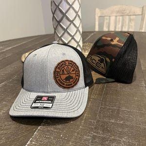 May include: Two trucker hats, one gray with a brown leather patch that says "The Dino Institute", and the other camouflage with a black mesh back.