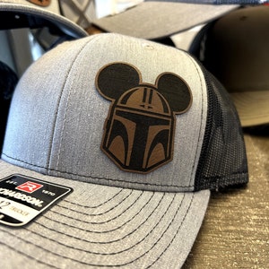 May include: A gray and black trucker hat with a brown leather patch featuring a Mickey Mouse silhouette with a Mandalorian helmet inside the ears. The hat has a black mesh back and a gray front with a black stitching detail. The hat has a red and white label with the text "Richardson 1970" on the front.