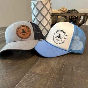May include: Two baseball caps with a black mesh back. The caps are gray and light blue. The front of each cap has a brown leather patch with the words "Hogwarts School of Witchcraft and Wizardry" and the Hogwarts crest.