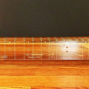 May include: A clear acrylic fret marker layout tool for a guitar neck, with the text "25.5 Scale" and "LBE Fret Marker Layout Tool". The tool is placed on a wooden guitar neck.
