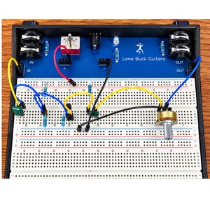 May include: A blue Lone Buck Guitars circuit board mounted in a black frame with a white breadboard. The setup includes various colored jumper wires, resistors, a metal potentiometer, a toggle switch, and input and output jacks for electronics.