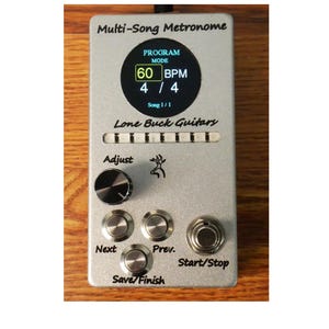 May include: A silver rectangular Multi-Song Metronome device by Lone Buck Guitars featuring a circular digital display showing 60 BPM and 4/4 time. The unit includes a black adjustment knob, four metallic push buttons labeled Next, Prev, and Start/Stop, and a Save/Finish button with a small deer logo.