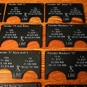 May include: A set of ten black plastic guitar nut templates with white text and a deer logo. Each template is labeled with a Fender guitar model name, such as "Fender '65" Strat "C"" and "Fender '57 Style Soft V". The templates are designed to help guitarists measure and adjust the nut on their guitars.