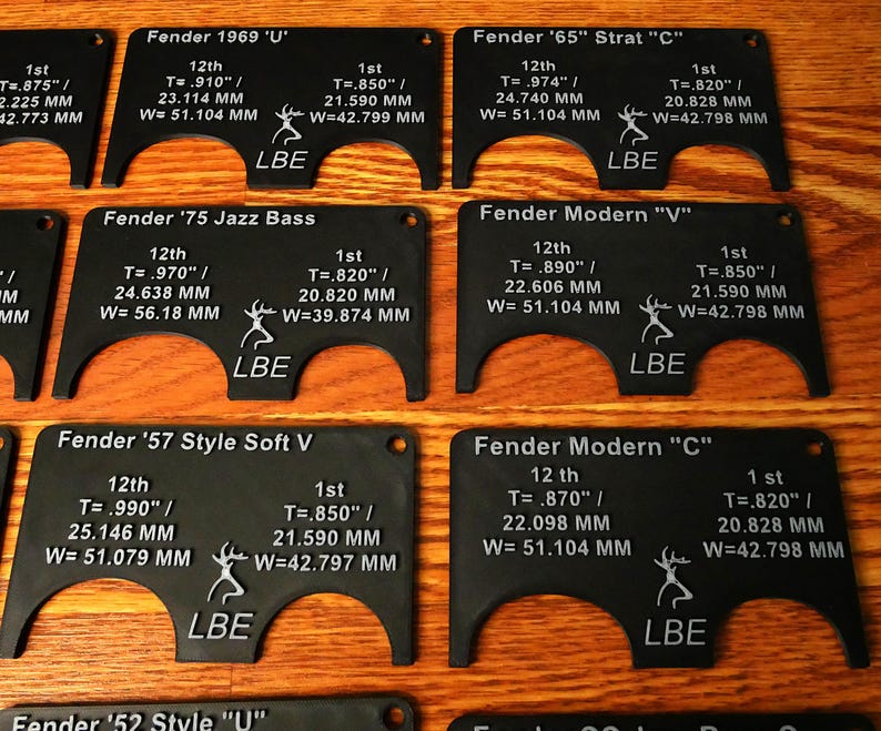May include: A set of ten black plastic guitar nut templates with white text and a deer logo. Each template is labeled with a Fender guitar model name, such as "Fender '65" Strat "C"" and "Fender '57 Style Soft V". The templates are designed to help guitarists measure and adjust the nut on their guitars.