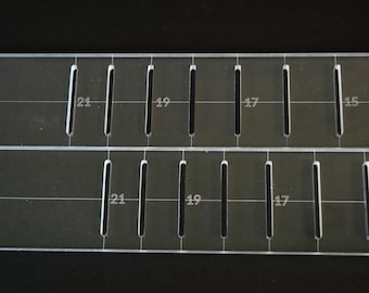 May include: Two clear acrylic rulers with evenly spaced vertical slots. Each ruler has numbers indicating measurements, including 15, 17, 19, and 21. The rulers are set against a dark background.
