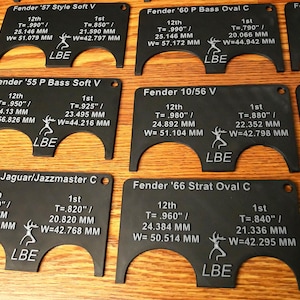 May include: A set of eight black plastic guitar nut measuring tools. Each tool is labeled with the guitar model, the 12th fret measurement in inches and millimeters, and the 1st fret measurement in inches and millimeters. The tools are labeled with the brand name "LBE".