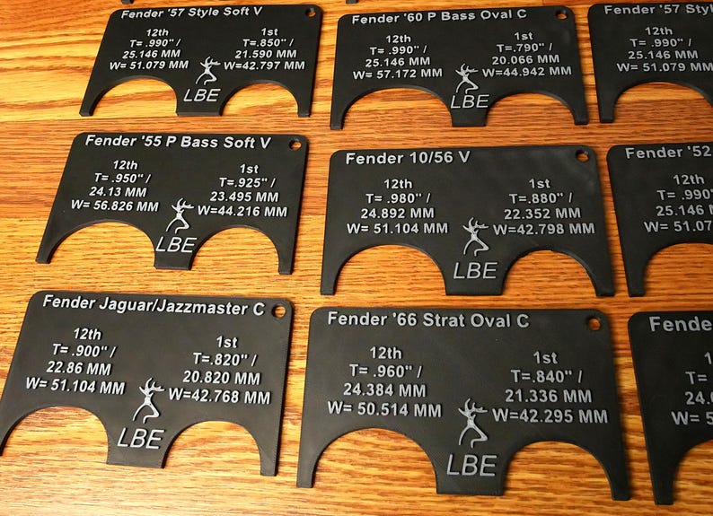 May include: A set of eight black plastic guitar nut measuring tools. Each tool is labeled with the guitar model, the 12th fret measurement in inches and millimeters, and the 1st fret measurement in inches and millimeters. The tools are labeled with the brand name "LBE".