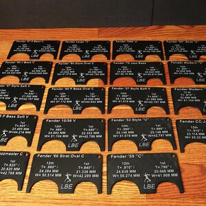 May include: A set of 14 black plastic templates with white text and markings. Each template is labeled with a Fender guitar model name, such as "Fender '57 Style Soft V" and "Fender '59 "C"". The templates are designed to help guitarists accurately measure and position their guitar parts.