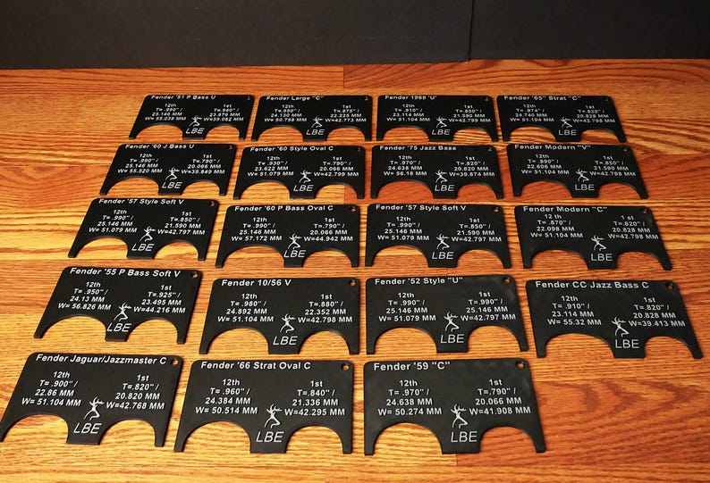 May include: A set of 14 black plastic templates with white text and markings. Each template is labeled with a Fender guitar model name, such as "Fender '57 Style Soft V" and "Fender '59 "C"". The templates are designed to help guitarists accurately measure and position their guitar parts.
