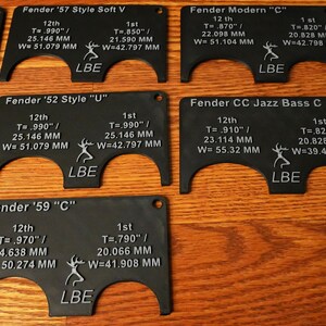May include: A set of six black plastic guitar nut measuring tools with white text and markings. Each tool is labeled with a Fender guitar model name and includes measurements in inches and millimeters. The tools are designed to help guitarists measure the correct nut width for their instrument.