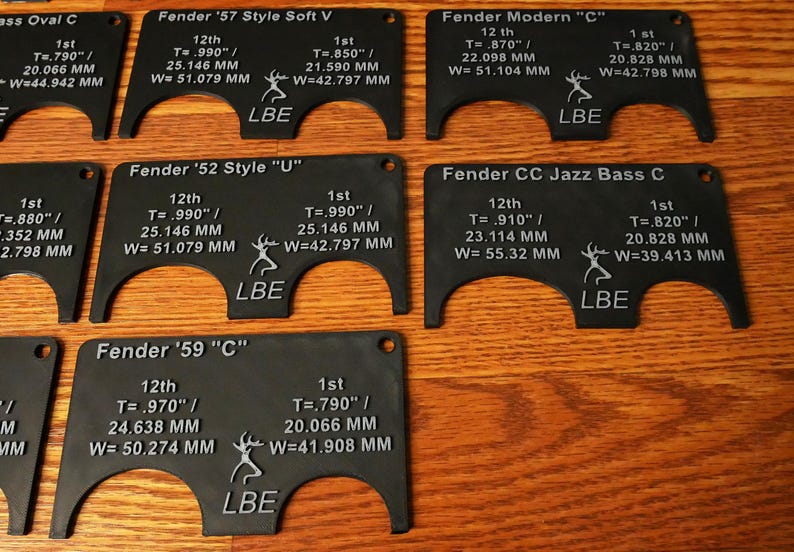 May include: A set of six black plastic guitar nut measuring tools with white text and markings. Each tool is labeled with a Fender guitar model name and includes measurements in inches and millimeters. The tools are designed to help guitarists measure the correct nut width for their instrument.