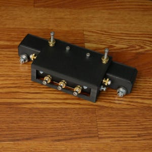 May include: A black plastic 3D printed device with four gold-colored wheels and two silver screws. The device has a black motor on the right side and a black plastic cover on the left side.