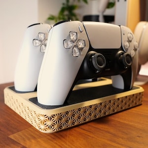 May include: A white and black gaming controller sits on a decorative wooden stand. The stand has a geometric pattern around the edges. The controller has a black centre section and white sides with black buttons.