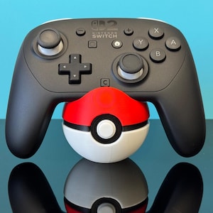 May include: A black Nintendo Switch Pro Controller sits atop a red, white, and black Poké Ball. The controller features a D-pad, buttons, and joysticks. The Poké Ball acts as a decorative stand.