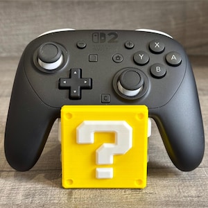 May include: A black Nintendo Switch Pro controller with white detailing, placed on a yellow block featuring a white question mark. The controller has buttons labelled X, Y, A, and B.