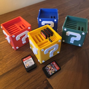 May include: A set of colourful, cube-shaped game cartridge holders. Each holder is a different colour: red, blue, green, and yellow. Each cube has a white question mark on two sides. Several game cartridges are visible.