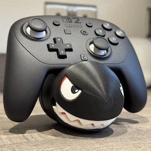 May include: A black Nintendo Switch Pro Controller with white buttons and a black, round, cartoon-like character attached. The character has a white mouth with teeth, a white eye, and a red eyebrow.