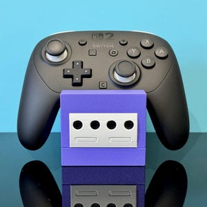 May include: A black Switch Pro controller with white detailing sits above a purple GameCube-style adapter. The adapter features a silver faceplate with four circular holes and two rectangular slots. The background is light blue.