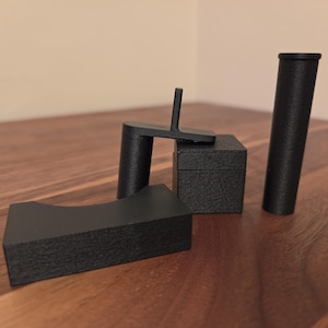 May include: A selection of black, geometric objects on a wooden surface. The objects include a curved rectangular piece, a small box, a cylindrical shape, and a piece with a perpendicular arm. The objects appear to be made of a textured material.
