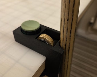 May include: A black 3D-printed accessory for a table, with a coin holder and a green button. A gold coin is visible in the holder, and the accessory is attached to a wooden leg. The table surface is white.