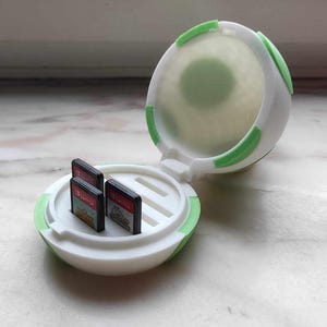 May include: A white and green spherical game cartridge holder, open to reveal three game cartridges. The holder has a hinged design with green accents. The cartridges are black with colourful game labels.