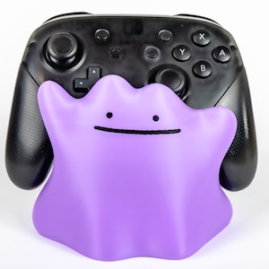 May include: A black Nintendo Switch Pro Controller with a purple Ditto-shaped accessory. The Ditto accessory has a simple face with a small smile and two dots for eyes. The controller has buttons labelled X, Y, A, and B.