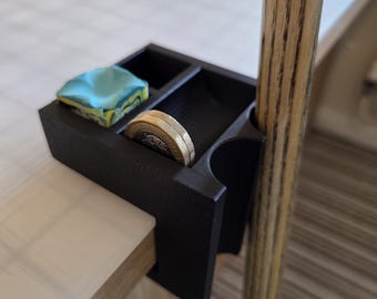 May include: A black pool table accessory, clamped to the table's edge, with compartments for chalk, coins, and a cue stick. It holds a blue and yellow chalk cube and coins. A wooden pool cue is visible.