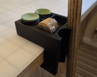 May include: A black pool table accessory holding two green circular pieces and a stack of coins. The accessory is attached to the side of a pool table and holds a pool cue.