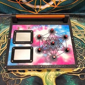 Może przedstawiać: Prostokątne urządzenie elektroniczne z uchwytem, ​​o kolorowym wzorze z motywem nieba. Posiada dwa prostokątne ekrany, wiele pokręteł z czerwonymi akcentami i geometryczny wzór. Urządzenie jest umieszczone na wzorzystym tle.