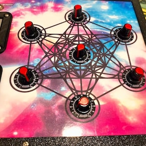 Może przedstawiać: Urządzenie elektroniczne z powierzchnią o tematyce galaktyki i geometrycznym wzorem. Posiada sześć czarnych pokręteł z czerwonymi wierzchołkami i ponumerowanymi tarczami. Urządzenie jest oprawione w czarną ramę.