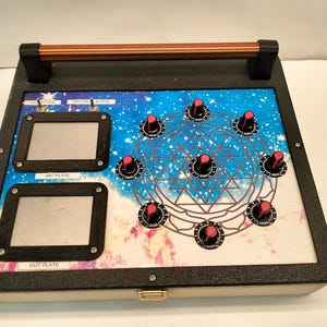 Może przedstawiać: Prostokątne urządzenie elektroniczne z czarną ramą, uchwytem i kolorową powierzchnią. Powierzchnia posiada dwa prostokątne ekrany, kilka pokręteł z czerwonymi osłonami i geometryczny wzór. Tło ma niebiesko-biały wzór przypominający galaktykę.