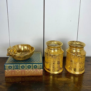 May include: Two gold metallic vases and a gold metallic bowl with a bird design on a stack of books. The vases and bowl have a textured surface.