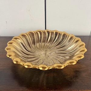 May include: A shallow, circular bowl with a metallic gold finish and a scalloped, wavy edge. The interior of the bowl features a radiating, fluted pattern that extends from the center toward the rim, creating a textured, reflective surface.