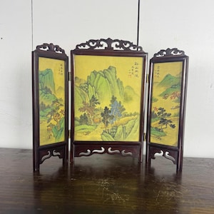 May include: A three-panel folding screen with a landscape painting. The screen has a dark wood frame and a yellow background. The painting depicts mountains, trees, and a waterfall. The screen is standing on a wooden surface.