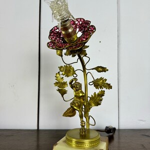 K&ouml;nnte beinhalten: Eine goldene Tischlampe mit einem rosa Blumenlampenschirm und einer Cherubfigur. Die Lampe steht auf einem Marmorsockel.