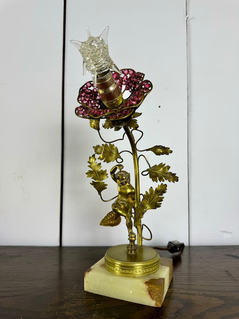 K&ouml;nnte beinhalten: Eine goldene Tischlampe mit einem rosa Blumenlampenschirm und einer Cherubfigur. Die Lampe steht auf einem Marmorsockel.