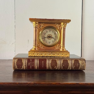 May include: A gold-toned mantel clock with a circular face, black numerals, and ornate column details, resting atop a brown leather-bound book titled The Essays of Elia by C. Lamb, featuring gold embossed lettering and decorative spine accents.